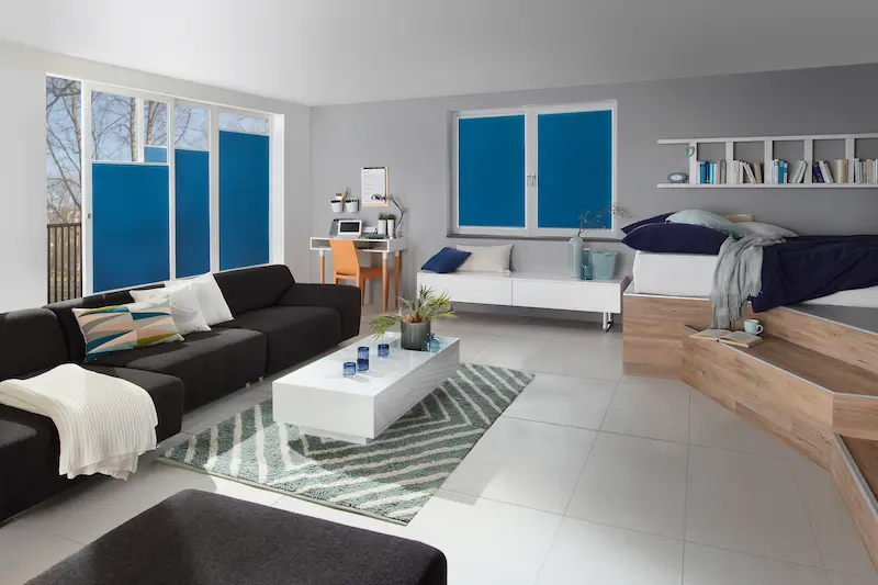 Modern eingerichteter Wohn und Schlafbereich mit blauen Wabenplissees als Sichtschutz an Fenstern und Tür